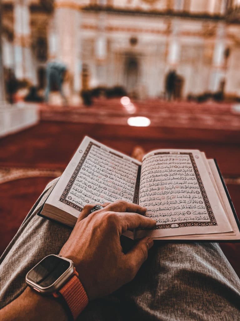 Student holding an open Mushaf practicing Quran recitation with measured pacing inside a mosque